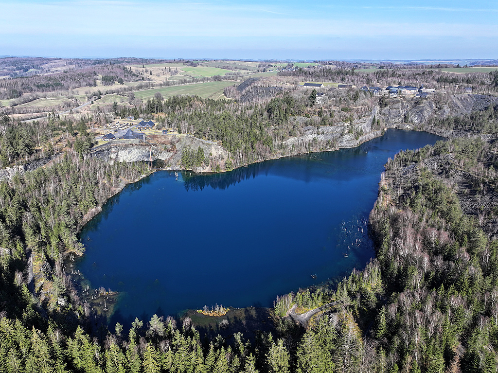 NSG 285, Blick von Südsüdwest auf den „Schiefersee“, gefluteter Abbaukessel des ehemaligen Schiefer-Tagebaus Kießlich, umgeben von Schieferwänden und -halden im Zentrum des NSG „Staatsbruch“ südsüdwestlich Lehesten (Landkreis Saalfeld–Rudolstadt). In der Bildmitte halblinks: das Museumsgelände. (Foto: Holm Wenzel, 09.03.2024)