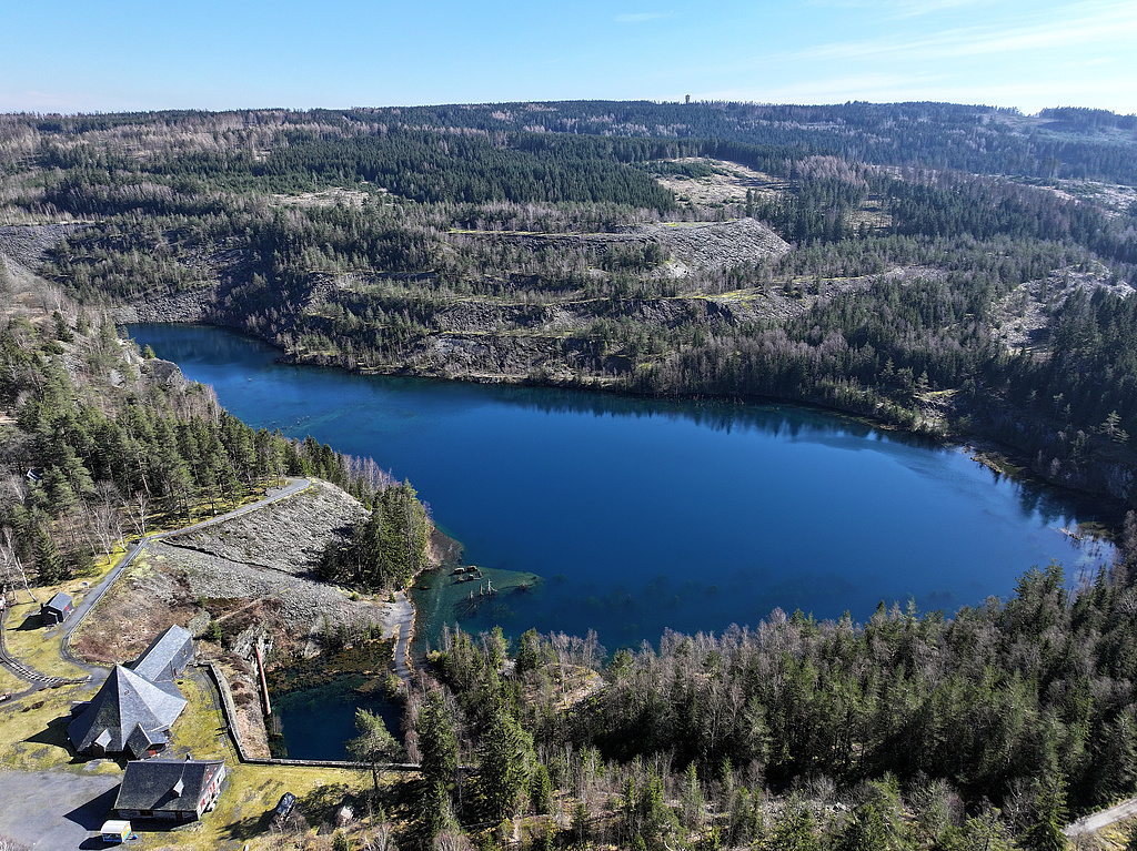NSG 285, Blick von Westnordwest auf den „Schiefersee“, gefluteter Abbaukessel des ehemaligen Schiefer-Tagebaus Kießlich, umgeben von Schieferwänden und -halden im Zentrum des NSG „Staatsbruch“ südsüdwestlich Lehesten (Landkreis Saalfeld–Rudolstadt). Vorn links: das Museumsgelände, ganz hinten: der Wetzstein, mit 792,7 m ü. NHN höchster Berg Ostthüringens und damit auch des Thüringer Teils des Frankenwaldes mit dem Altvaterturm. (Foto: Holm Wenzel, 09.03.2024)