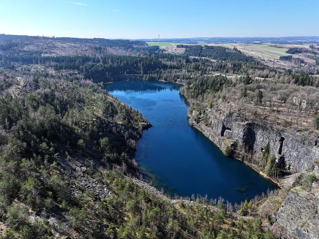 NSG 285, Blick von Nordosten auf den „Schiefersee“, gefluteter Abbaukessel des ehemaligen Schiefer-Tagebaus Kießlich, umgeben von Schieferwänden und -halden im Zentrum des NSG „Staatsbruch“ südsüdwestlich Lehesten (Landkreis Saalfeld–Rudolstadt). In der Bildmitte halbrechts: das Museumsgelände. (Foto: Holm Wenzel, 09.03.2024)
